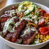 Charred roasted corn and tender sliced steak top a vibrant bowl with avocado slices and zesty cilantro cream sauce.